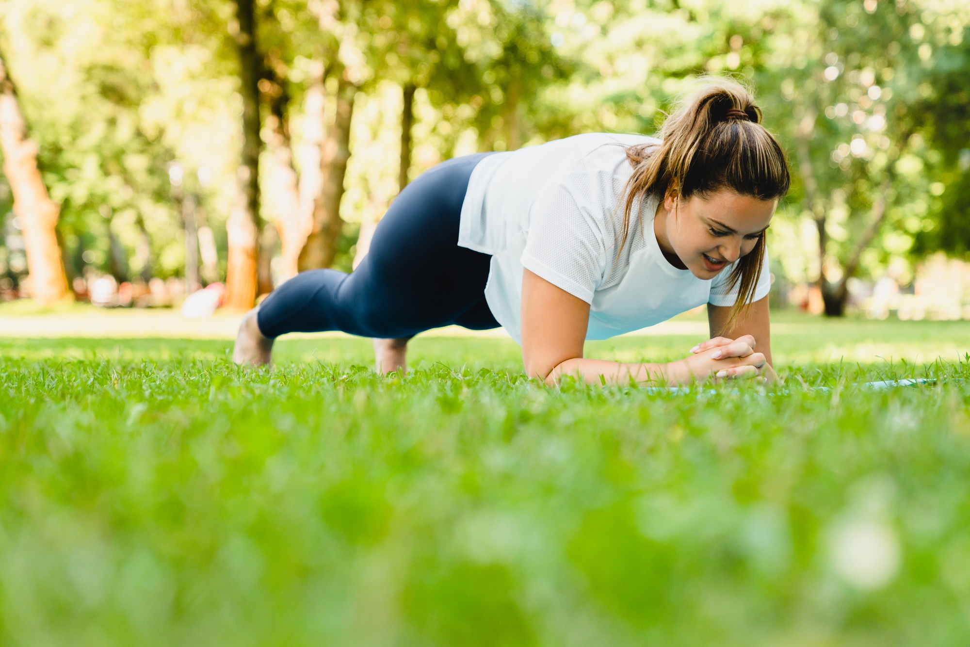 Athlete standing in plank position, losing weight on fitness yoga training class on lawn in park